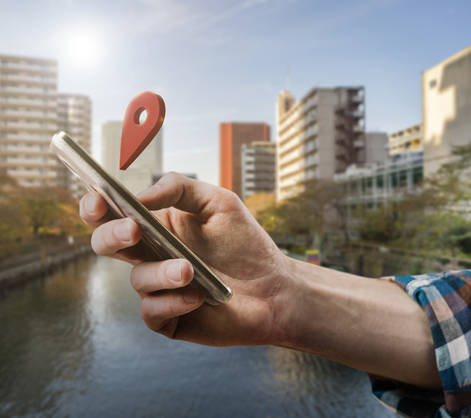 Imagem de uma mão segurando um smartphone, com um pino de localização vermelho 3D flutuando sobre ele, em um cenário urbano com um rio. O contexto visual remete à Geolocalização e SEO Local, representando a conexão de negócios com sua comunidade. O foco é para quem busca palestrante sobre como posicionar empresas como referência local e aumentar o engajamento na região.